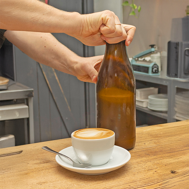 eine Flasche Haferdrink und eine Tasse Cappuchino mit Latte Art Herz